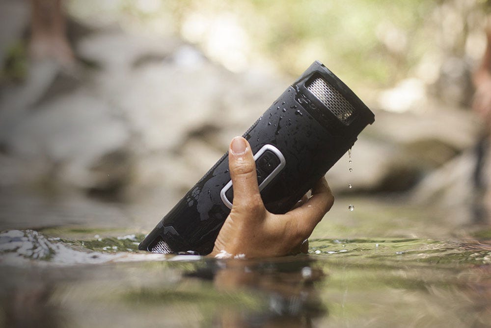 man holding wireless portable speaker above water in nature setting