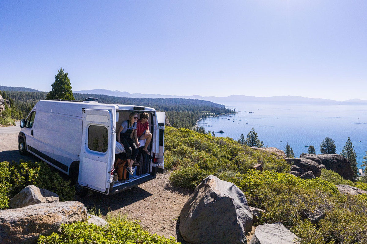 Two people sitting in white camper van overlooking forest and lake