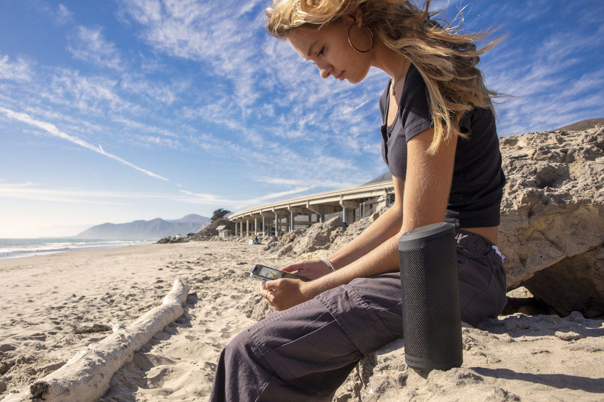 woman sitting beach listening to music via Scosche wireless speaker