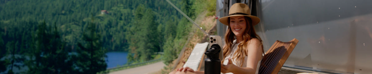 Woman with a hat on sitting by a camper looking at her phone attached to a water bottle