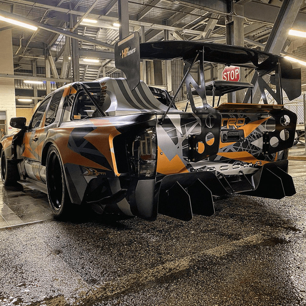 A custom Toyota Tacoma race truck sits in a garage with orange and black graphics and a spoiler installed.