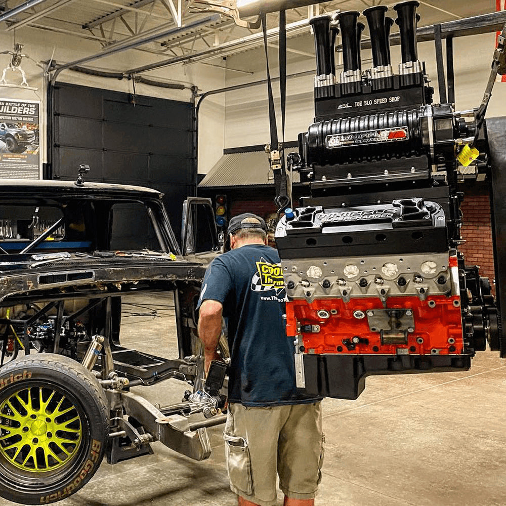 A large race engine hangs in a garage next to a truck frame and a professional installer.