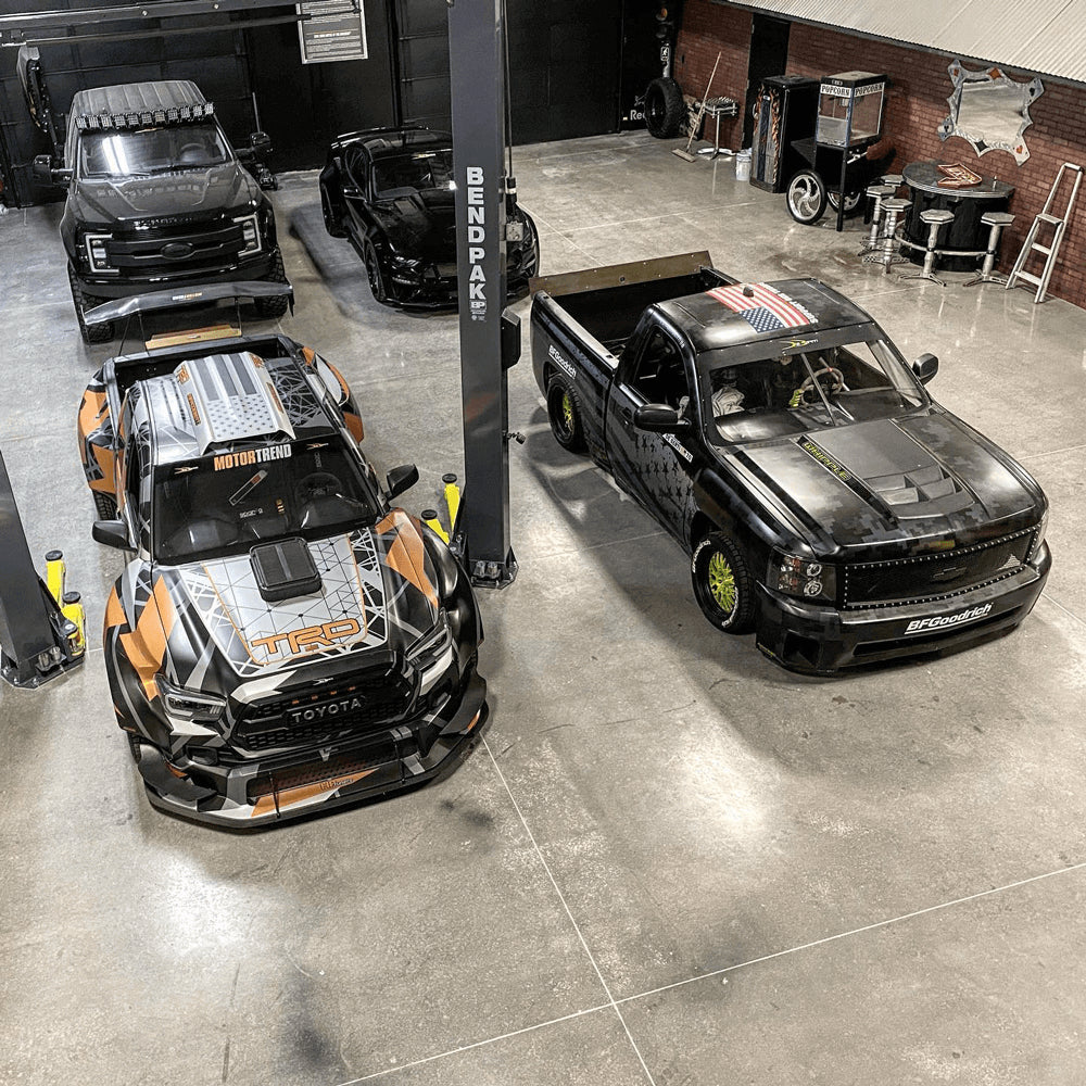 Four customized vehicles sit in a garage, including a large black Ford truck, a Tacoma Tacoma race-car, a black Chevy race-truck and small black sports-car.