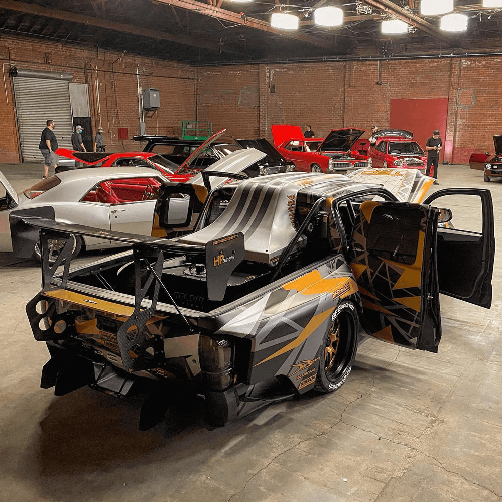 A custom Toyota Tacoma race truck with orange and black graphics sits in a garage, next to six classic show-cars.