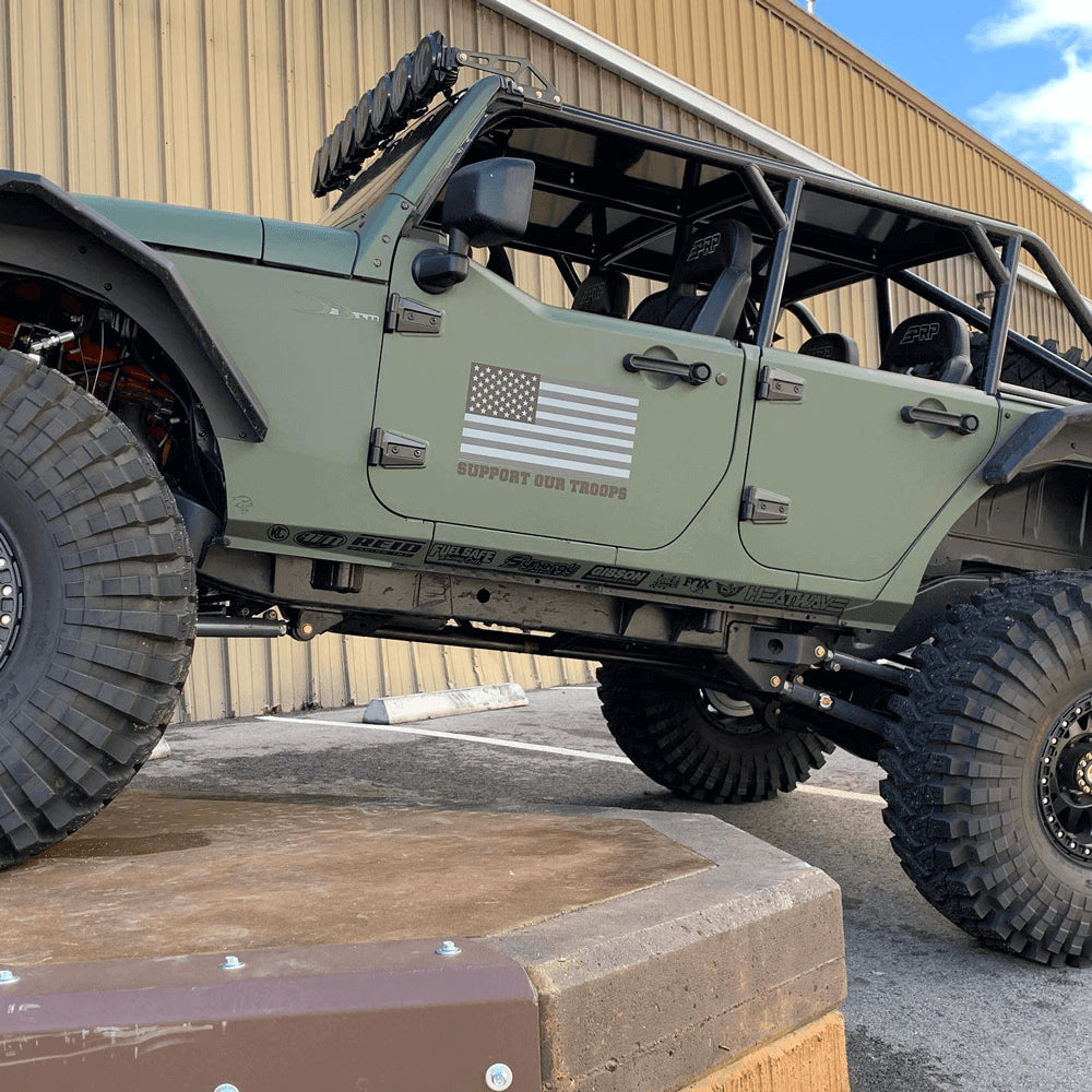 A customized matte-green Jeep with an American flag on the door that reads Support The Troops, is perched on a high curb showing off-road capabilities.