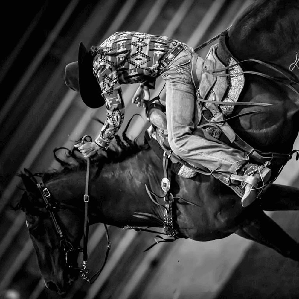 A cowgirls rides a dark horse in a rodeo and holds on tightly to the reigns.