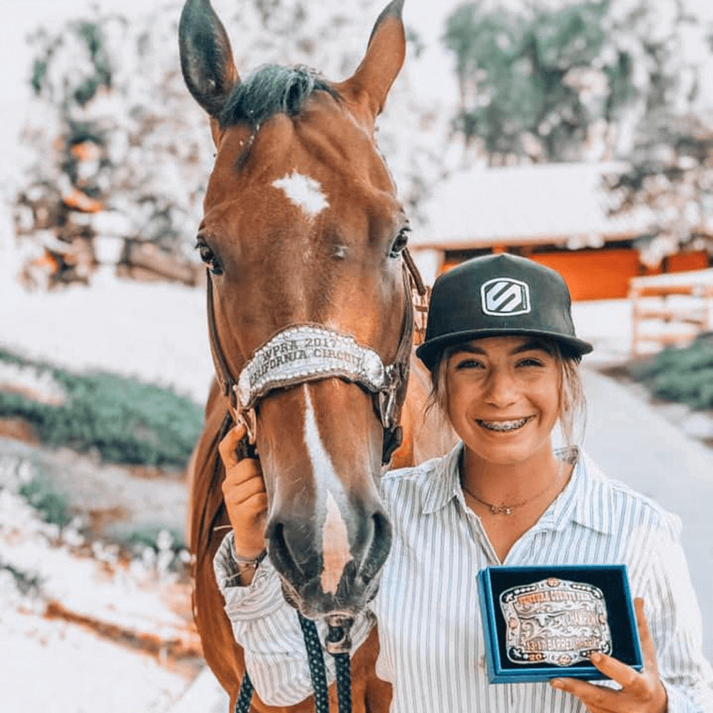 Skyler Alves poses with her horse, she's holding a belt buckle and there's snow, trees and a barn in the background.