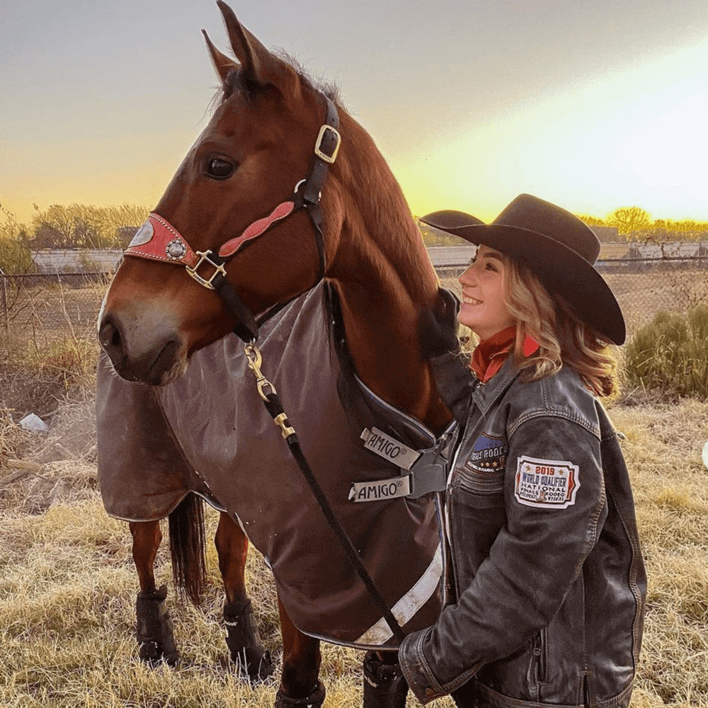 Skyler Alves poses with her horse, both are wearing coats and there is a sunset in the background.