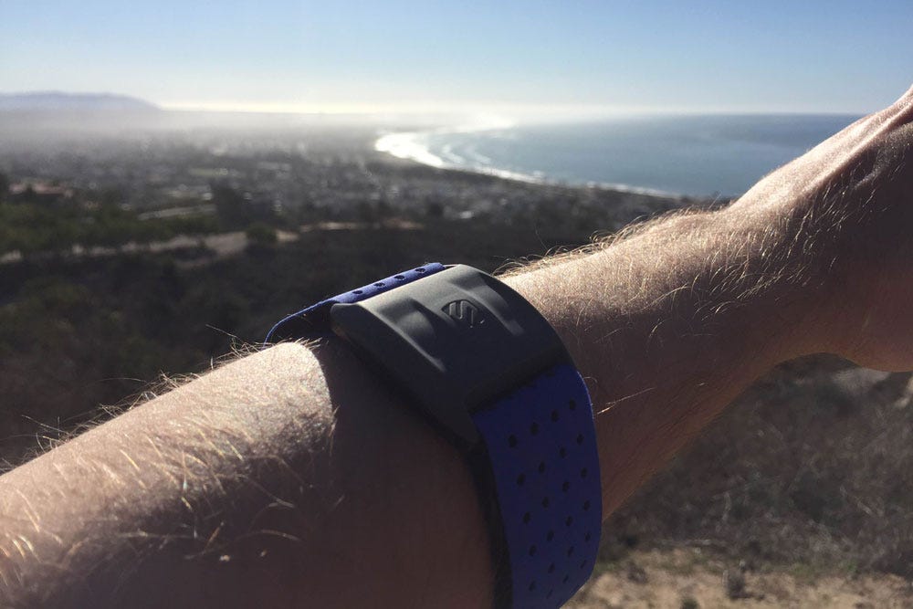 close up of man's arm wearing Scosche heart rate monitor with ocean and city in background