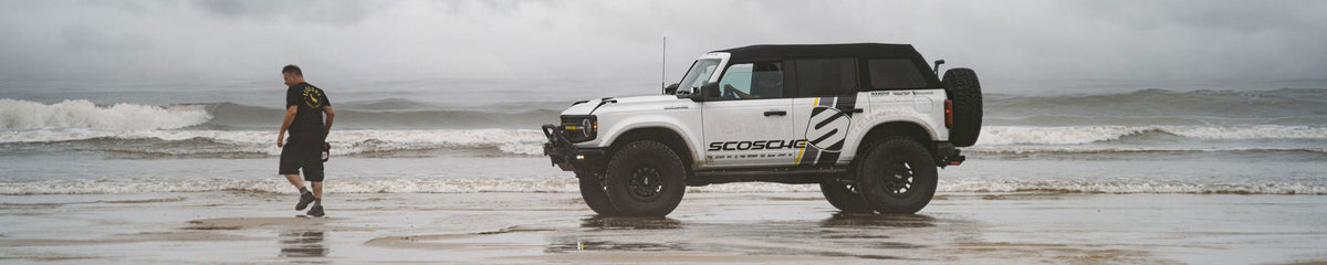 White Ford Bronco on beach with man standing next to it holding a camera with waves crashing behind