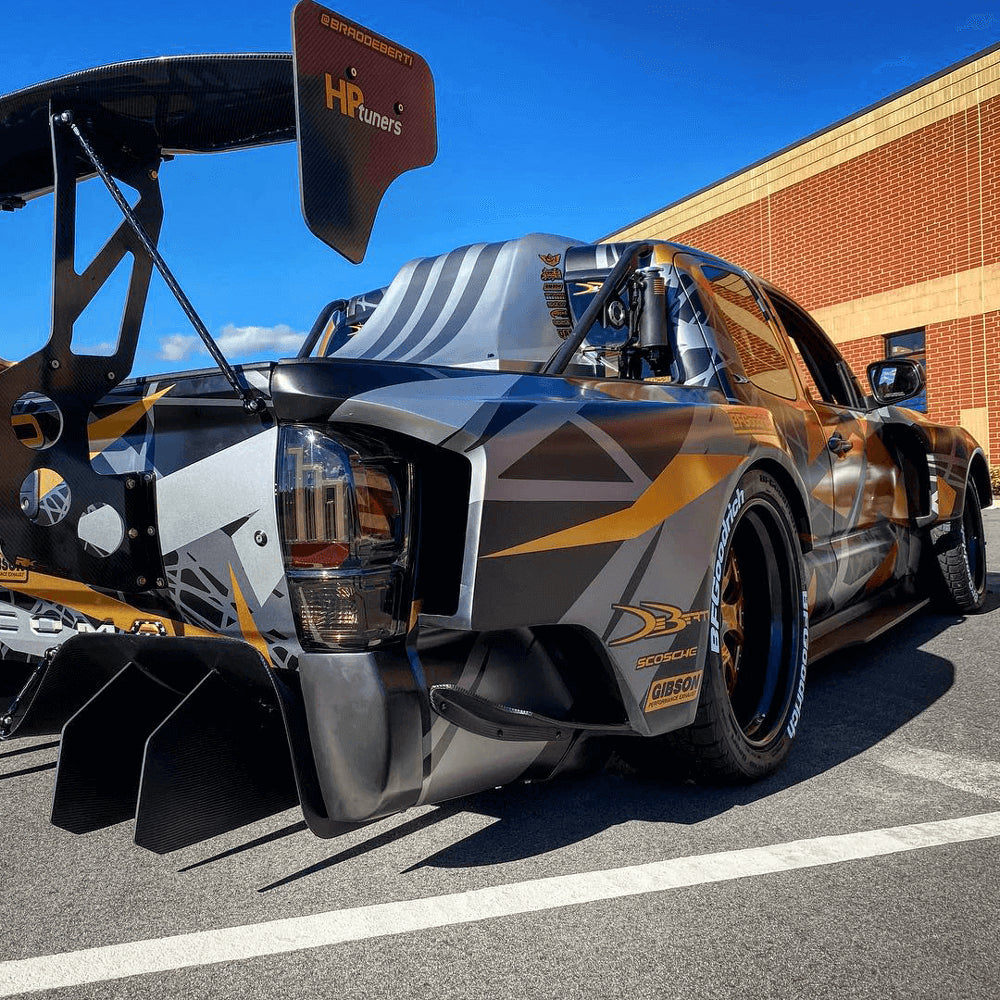 A custom Toyota Tacoma race truck sits in a parking lot with orange and black graphics and a spoiler installed.