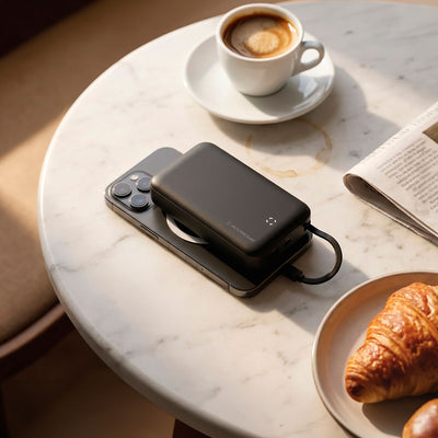 Phone on table top with a power bank on top of it plugged in with a coffee and crossant on the table