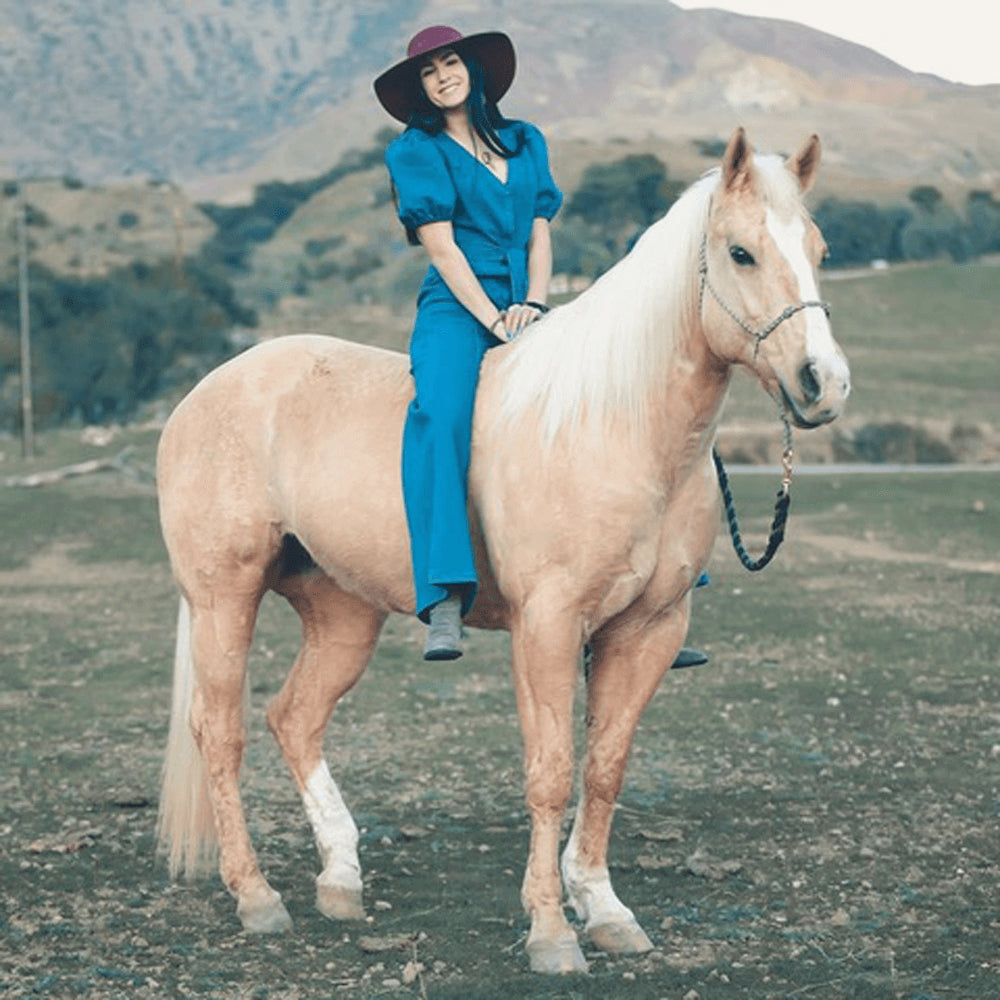 Pfieffer is wearing a blue outfit and sits on a muscular tan horse with hills and trees in the background.