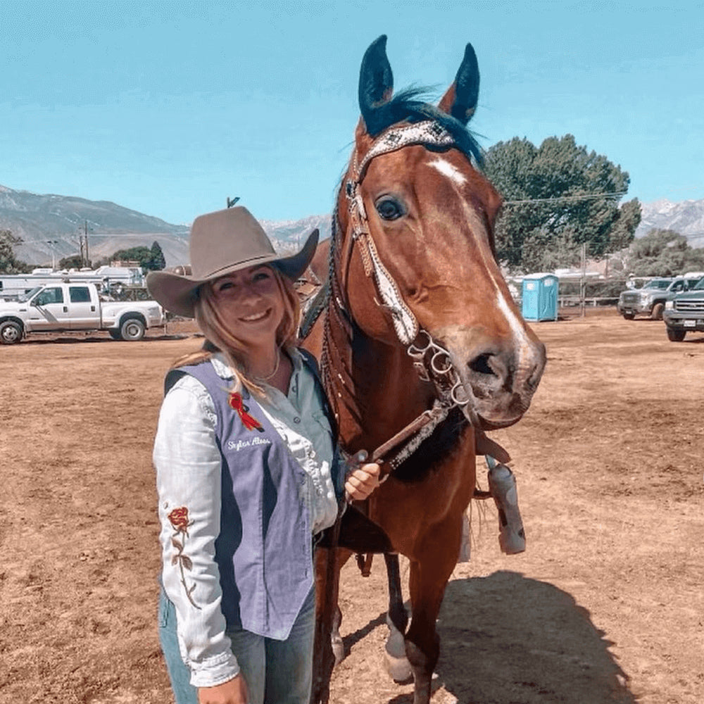Skyler Alves poses with her horse with trucks, trees and hills in the background.