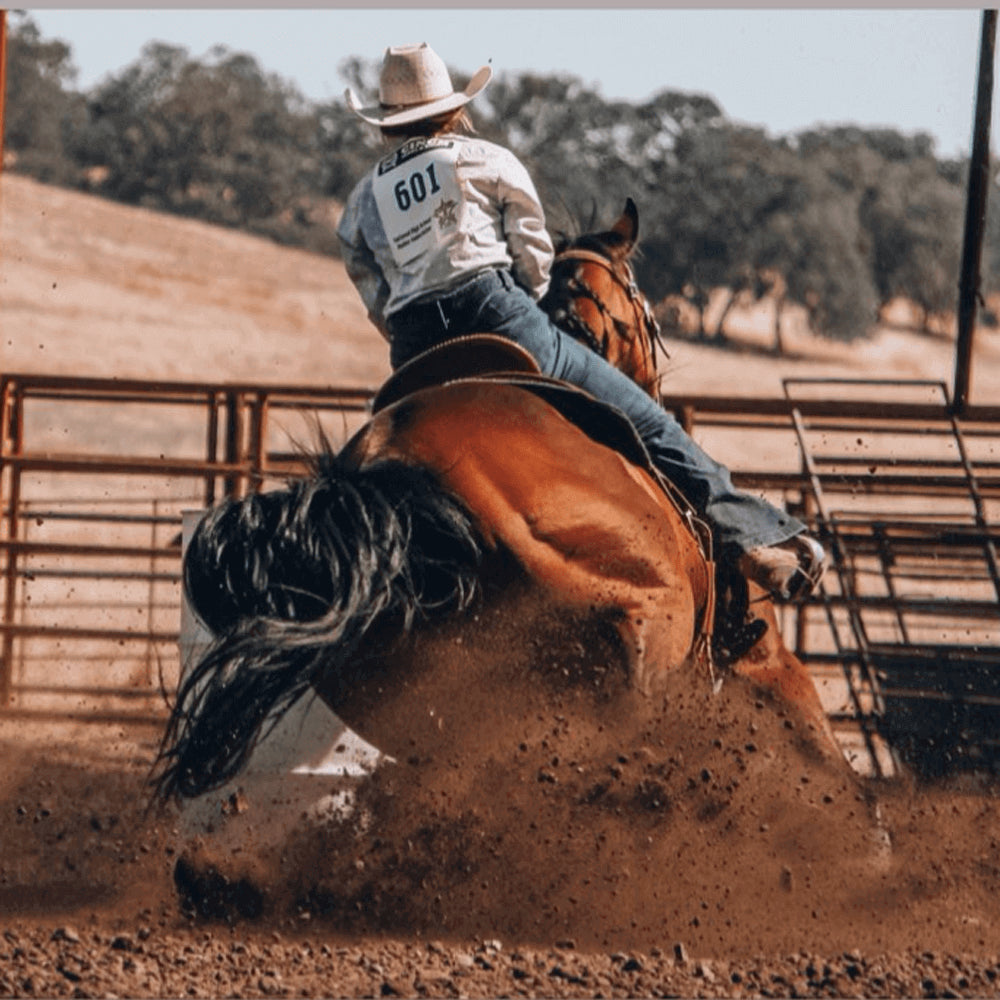 A cowgirl rides a horse aggressively in a rodeo with hill and trees in the background.