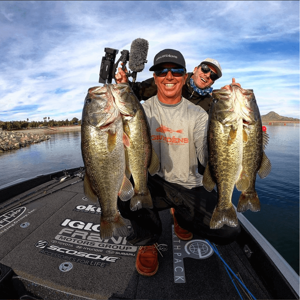 Man kneeling holding four fish with another man behind him holding a camera