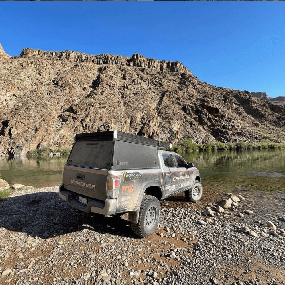 A dirty custom Toyota Tacoma sits next to a river, with rocky cliffs behind it